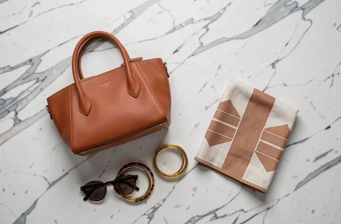 Flat lay d'accessoires de mode tendance : sac en cuir cognac, bracelets dorés, lunettes et foulard en lin sur marbre blanc