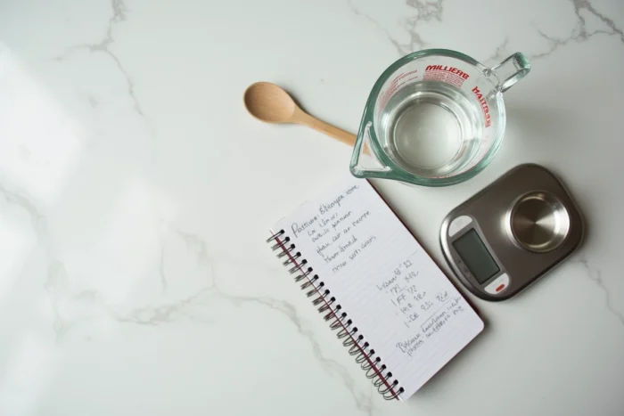 Vue rapprochée d'un verre doseur, d'une balance numérique et d'un carnet de conversion grammes en litres sur un plan de travail en marbre
